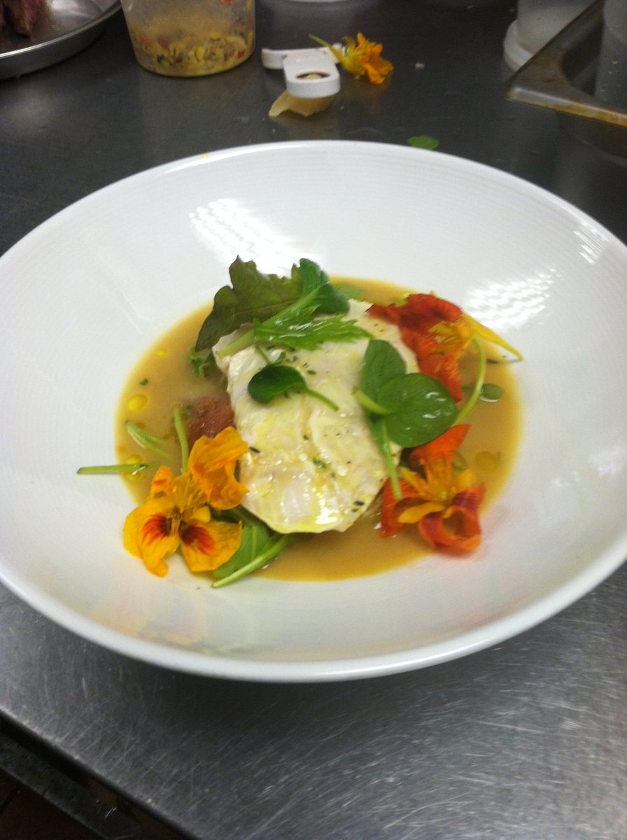 White fish in broth with greens and nasturtium flowers in a white bowl