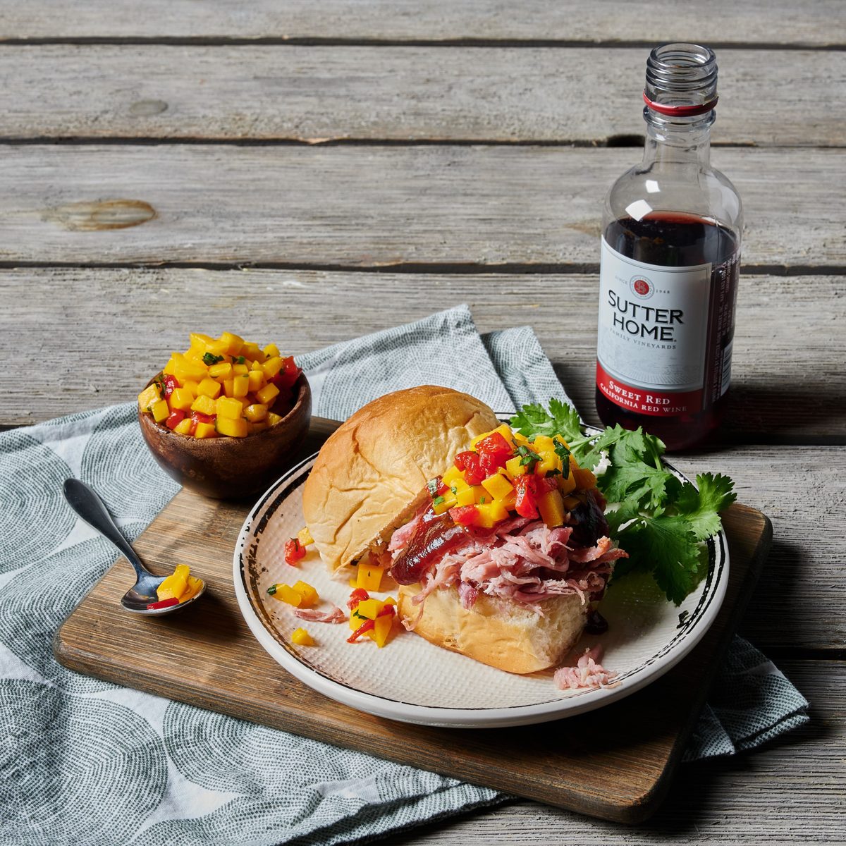 Pulled pork sandwich topped with mango salsa beside a mini bottle of sweet red wine