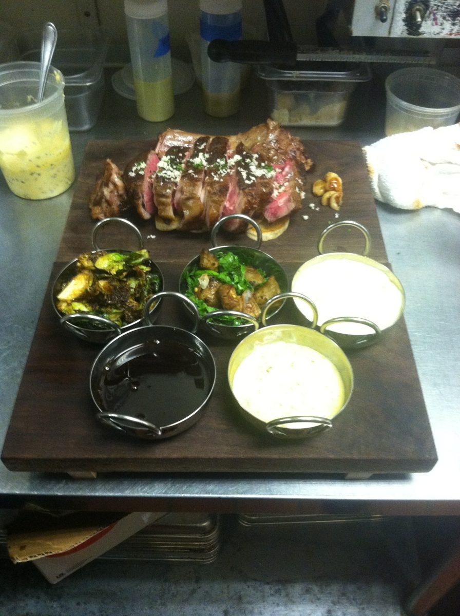 Sliced ribeye on a wooden board with sides and sauces in small copper pans