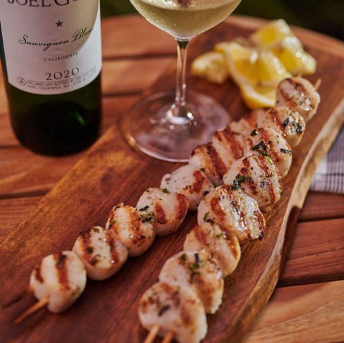 Grilled scallop skewers on a wooden board with lemon and a glass of white wine